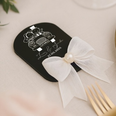 Lembrança de casamento preta com gravura branca de noivos em carro, laço branco com pérola, sobre mesa com talheres dourados e prato.