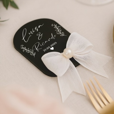 Cartão decorativo preto com texto branco e laço branco com pérola, em cima de tecido bege, ao lado de talheres dourados.