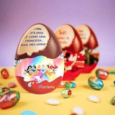 Ovo de chocolate com imagem de princesas Disney e texto em português sobre superfície amarela com pequenos ovos coloridos