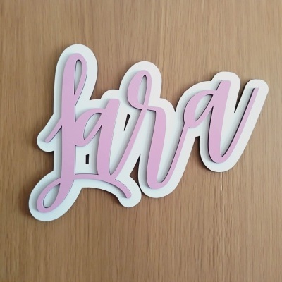 Placa decorativa com a palavra Lara em rosa e branco sobre fundo de madeira.