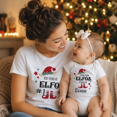 Mãe e bebé com t-shirts brancas com estampados de elfos, sentados num sofá junto a uma árvore de Natal iluminada.