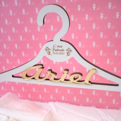 Cabide branco em madeira com texto dourado em fundo rosa com padrão religioso