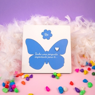 Cartão com borboleta azul, penas brancas e pedras coloridas sobre superfície rosa