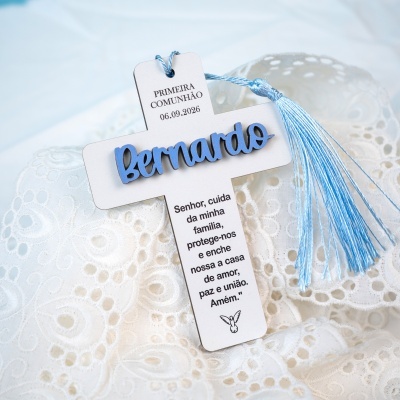 Cruz branca decorativa com nome Bernardo em relevo azul e franja azul sobre tecido branco bordado