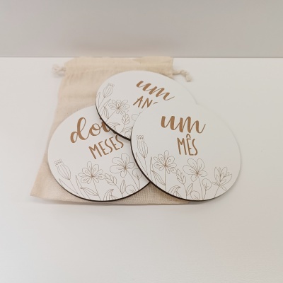 Placas redondas em madeira com frases em castanho e desenhos de flores com saco de tecido.