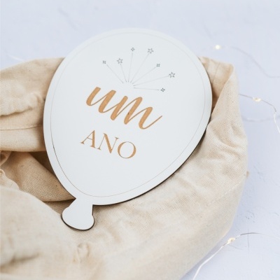 Placa decorativa de balão branco com texto dourado um ANO sobre tecido bege e fundo claro
