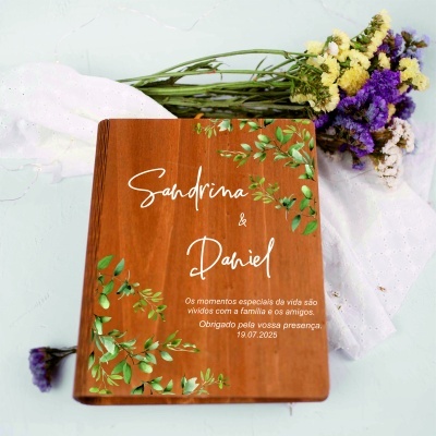 Livro de madeira decorado com texto branco e folhas verdes, junto a flores coloridas e tecido branco em fundo claro.