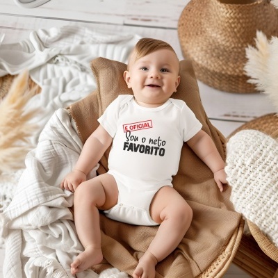 Bebé sorridente com body branco e texto impresso sentado numa almofada castanha e fundo com tecidos brancos e saco de ráfia.