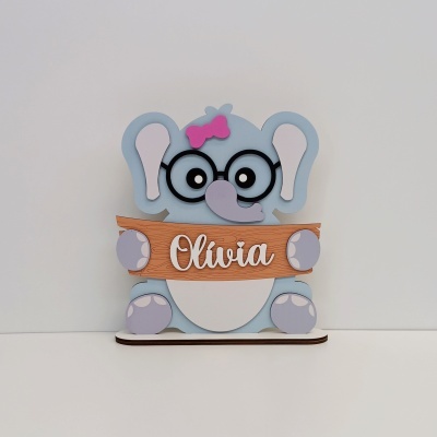 Figura de elefante azul com laço cor-de-rosa segurando placa com nome Olívía