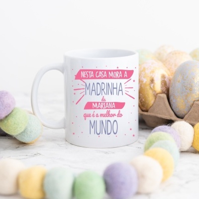 Caneca branca personalizada com texto para madrinha rodeada por ovos decorativos e guirlanda colorida.