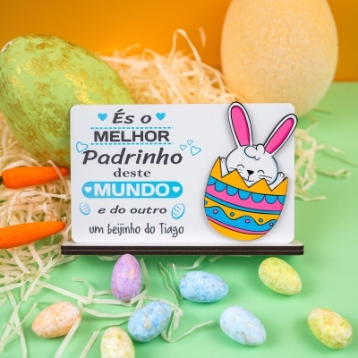 Placa decorativa com texto em português e coelho colorido, ovos e cenouras decorativas ao fundo