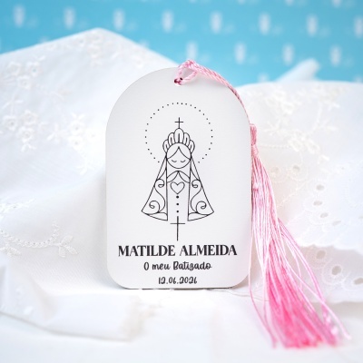 Etiqueta de batizado branca com desenho religioso e fio rosa, sobre tecido branco bordado