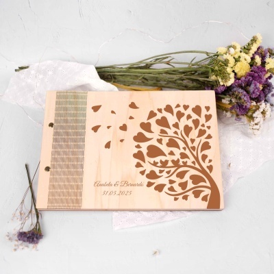 Livro de capa em madeira com desenho de árvore de corações e texto, ao lado de flores secas sobre fundo claro.