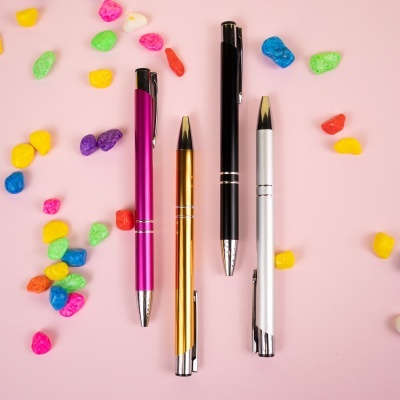 Quatro canetas metálicas coloridas sobre fundo rosa com pedras decorativas multicoloridas