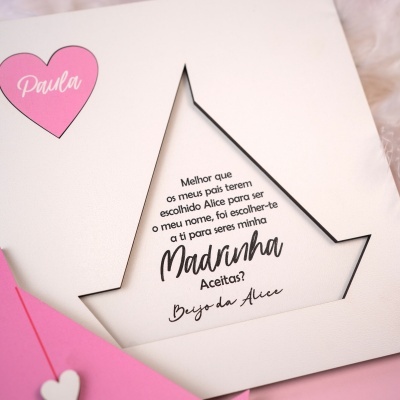 Cartão branco com recorte de estrela e coração rosa com nome Paula sobre envelope rosa em fundo de tecido macio