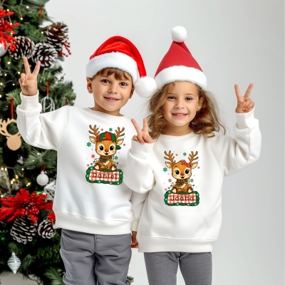 Crianças com sweatshirts brancas de Natal com nome e desenhos de renas, usando gorros de Natal vermelhos e brancos perante árvore decorada