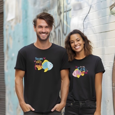 Homem e mulher sorrindo com t-shirts pretas com frases e ilustrações de peixes, parede grafitada ao fundo.