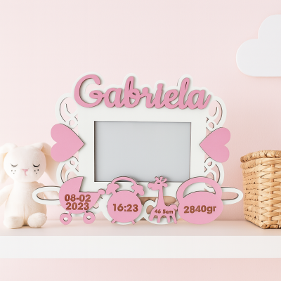 Moldura porta-retrato rosa e branca com nome Gabriela e símbolos de nascimento sobre prateleira com peluche e cesta