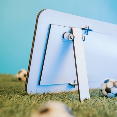 Parte traseira de um espelho com suporte em madeira clara sobre relva artificial e bolas de futebol ao fundo