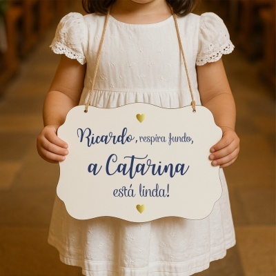 Placa branca de madeira com texto em azul e dourado, segurada por criança com vestido branco bordado