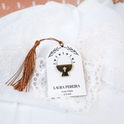 Medalha dourada com cruz num cartão branco com texto, sobre tecido branco bordado