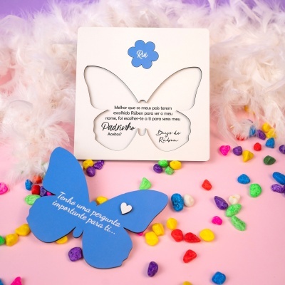 Cartão convite borboleta branco e azul com texto em português rodeado por penas brancas e pedras coloridas sobre superfície rosa