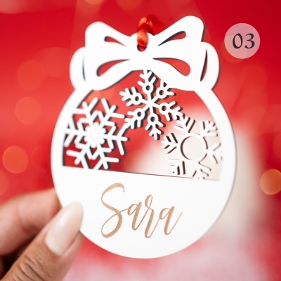 Etiqueta branca com laço e flocos de neve com fio vermelho e texto Sara em dourado