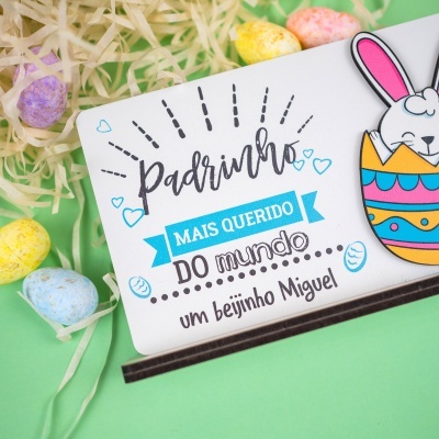 Caixa branca com autocolante de coelho colorido e texto de Páscoa, acompanhada de ovos coloridos em fundo verde.