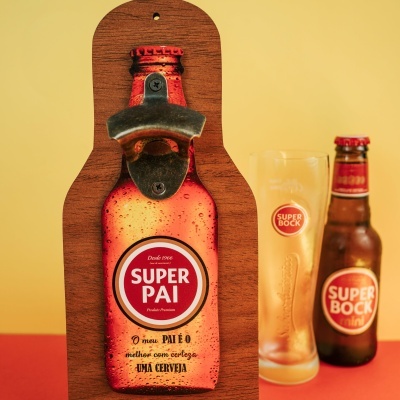Abridor de garrafas em metal numa base de madeira com imagem de garrafa de cerveja e texto SUPER PAI e copo e garrafa de cerveja Super Bock mini ao fundo