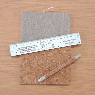 Caderno com capa bege e base de cortiça com régua branca e lápis de cortiça sobre mesa de madeira.