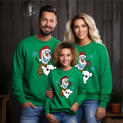 Três pessoas com sweatshirts verdes com boneco de neve Olaf, em frente a parede de madeira escura com lâmpada pendurada e plantas em vasos.