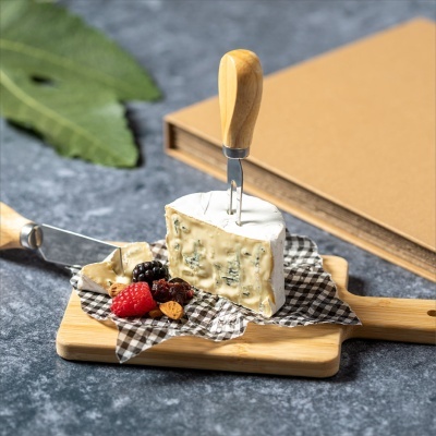 Queijo cremoso com nódoas azuis numa tábua de madeira com frutos e utensílios com cabos de madeira.