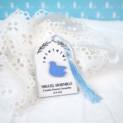Etiqueta branca com pendente azul e tassel azul claro sobre tecido branco e renda, fundo azul e branco.