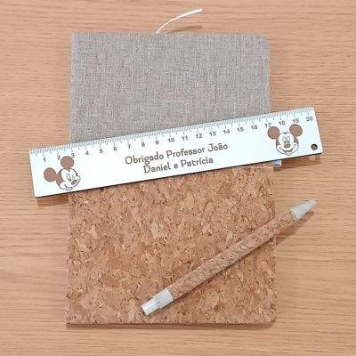 Régua branca com o texto Obrigado Professor João Daniel e Patrícia, caderno cinzento, bloco de cortiça e caneta revestida de cortiça.