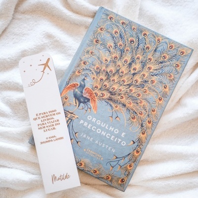 Livro Orgulho e Preconceito com capa azul e ilustrada e marcador de livros branco deitado sobre tecido branco