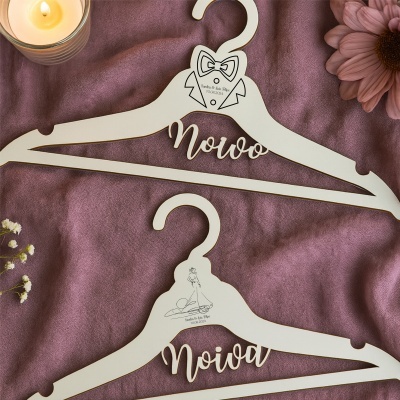 Cabides de madeira creme personalizados para noivo e noiva sobre pano púrpura com vela acesa e flor rosa