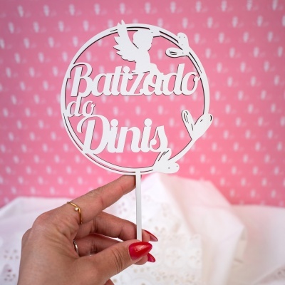 Decoração em madeira branca com texto Batizado do Dinis e anjo, segurada por mão feminina num fundo rosa com padrão branco.