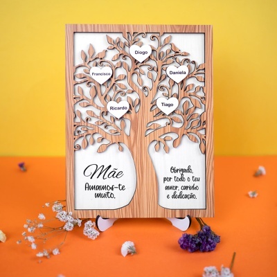 Quadro decorativo de madeira com árvore recortada e nomes em corações, texto em português dedicado à mãe, fundo amarelo e superfície laranja com flores.