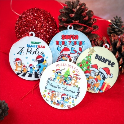 Enfeites de Natal redondos personalizados com desenhos coloridos e texto, dispostos sobre fundo vermelho com pinhas e esfera vermelha brilhante.