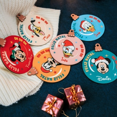 Etiquetas de Natal Disney coloridas com nomes, sobre tecidos branco e azul e dois pequenos presentes rosa.