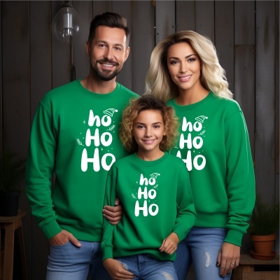 Três pessoas com sweatshirts verdes com texto branco ho Ho Ho, calças de ganga, fundo de madeira escura e planta em vaso.