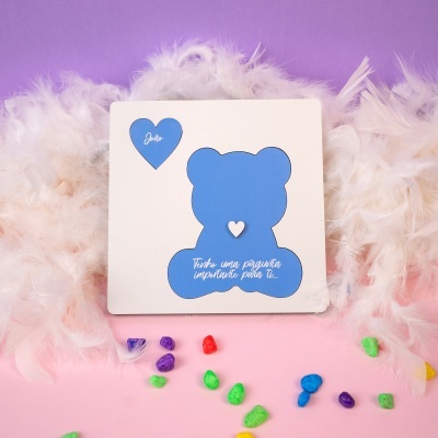 Cartão branco com desenho azul de urso e coração com texto João, penas brancas e pedras coloridas em fundo rosa e roxo
