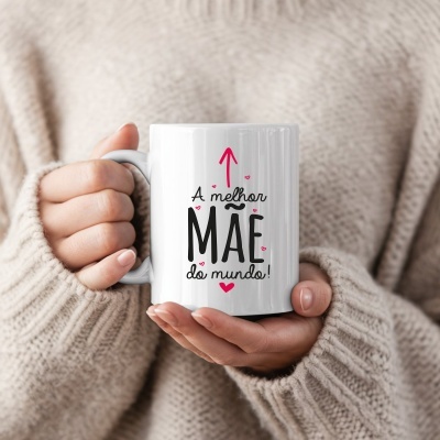 Caneca branca com texto A melhor MÃE do mundo segurada por mãos e suéter bege