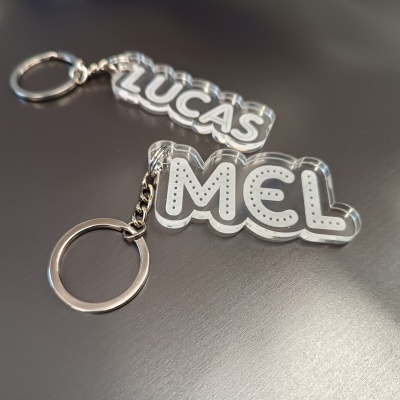 Chaveiros transparentes com nomes LUCAS e MEL em letras brancas.
