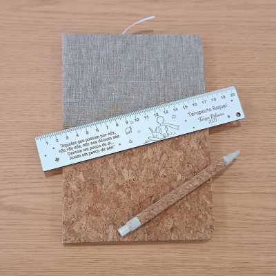 Caderno com capa de tecido cinzento, régua branca com texto e caneta revestida a cortiça sobre mesa de madeira.
