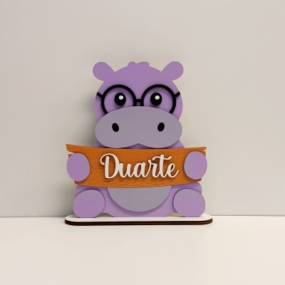 Boneco decorativo de hipopótamo roxo com óculos segurando placa de madeira com o nome Duarte