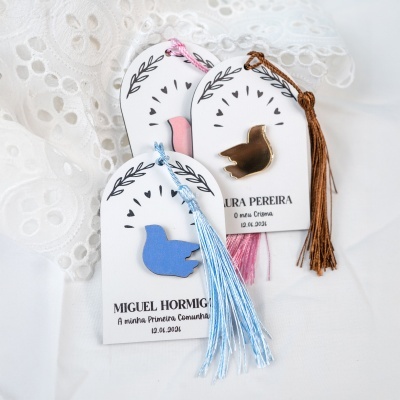 Lembranças decorativas com pombas em azul, rosa e dourado e fios pendurados sobre fundo de tecido branco com renda