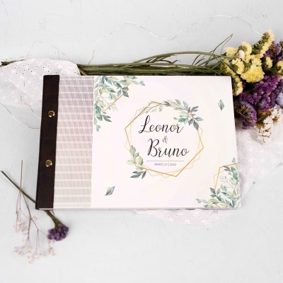Álbum de casamento branco com detalhes de folhas verdes e nomes Leonor & Bruno, rodeado por flores secas coloridas.