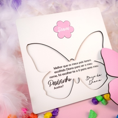 Cartão de borboleta em papel branco com texto e flor rosa com nome Diana, sobre fundo de penas e objetos coloridos