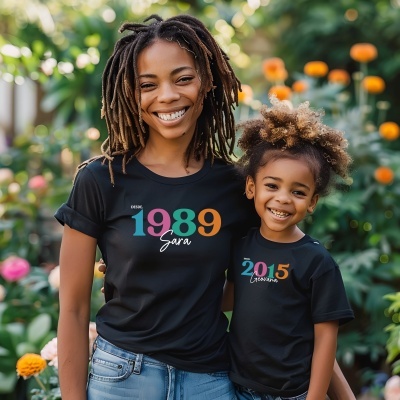 Duas pessoas com t-shirts negras com estampas coloridas e nomes, em ambiente com plantas e flores.
